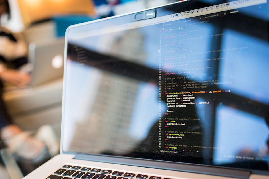 A close-up of a laptop screen displaying lines of colorful programming code with syntax highlighting, set in a bright workspace with blurred background elements such as another laptop and a person wor