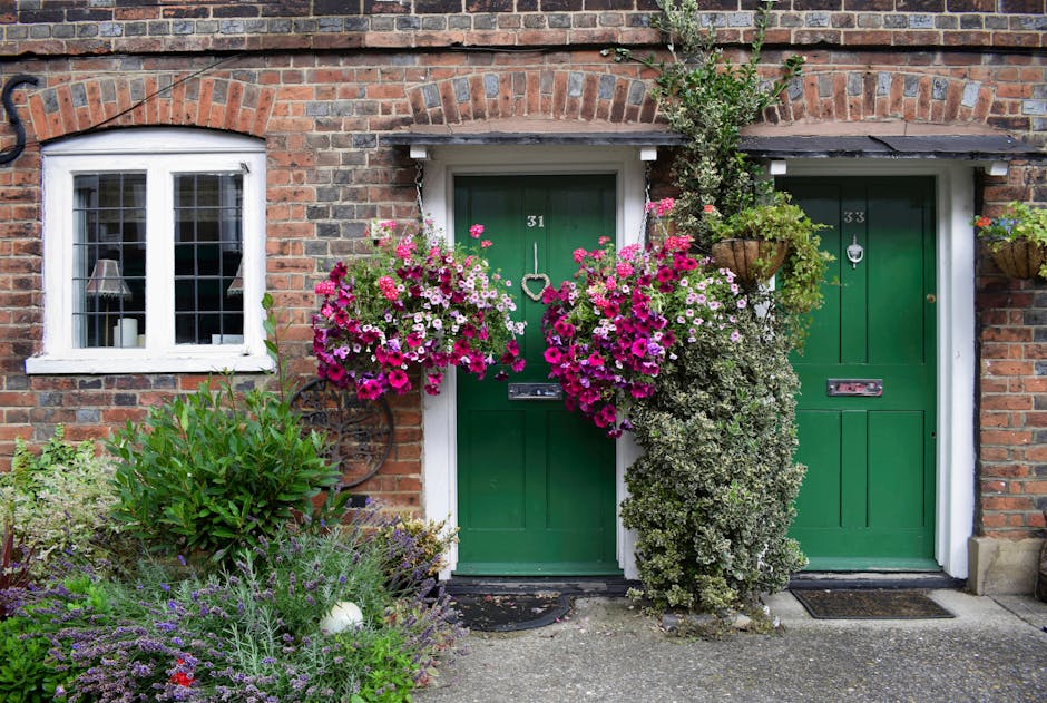 A charming residential brick property featuring two green front doors, numbered 31 and 33, each adorned with metallic mail slots and door knockers. The area showcases well-maintained garden beds in fr