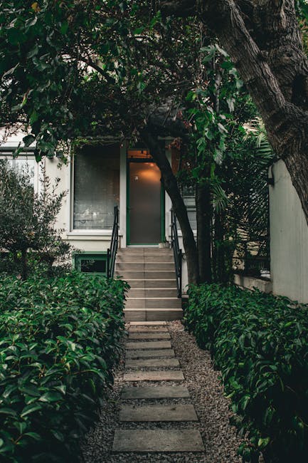 A narrow garden pathway leading to a house entrance, bordered on both sides by lush, well-maintained green shrubbery and foliage. The path consists of rectangular stone stepping stones set on gravel, 